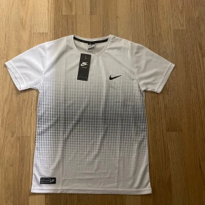 Vit t-shirt från Nike - Säljer en snygg vit t-shirt från Nike i storlek M. Den har ett coolt svart mönster som tonar ut nedåt och en liten Nike-logga på bröstet. Perfekt för träning eller vardagsbruk. Materialet är lätt och andas bra. Passa på att köpa denna stilrena t-shirt! 🖤