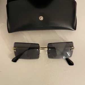 Svarta rektangulära solglasögon med gulddetaljer - Snygga svarta rektangulära solglasögon med gulddetaljer och en stilren design. Perfekta för en cool look året runt. Kommer med ett elegant svart fodral för säker förvaring.