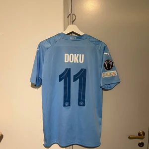 Ljusblå Manchester City fotbollströja - Säljer en ljusblå Manchester City fotbollströja med Doku och nummer 11 på ryggen. Tröjan är kortärmad och har klubbens emblem samt Puma-loggan på framsidan. Den har också ett märke på ärmen. Perfekt för fans som vill visa sitt stöd! Storlek M! Hör av er vid några frågor!
