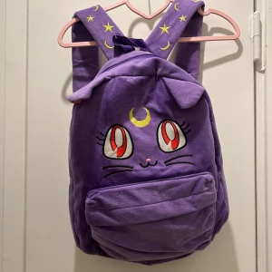 Lila ryggsäck med kattmotiv sailor moon - Supergullig lila ryggsäck med ett broderat kattansikte och måne på framsidan, sailor moon motiv. Den har justerbara axelband med stjärnor och månar. Perfekt för skolan eller som en söt accessoar. Ryggsäcken har en praktisk framficka med dragkedja.