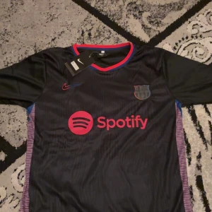 Svart FC Barcelona tröja med Pedri 8 - Säljer en svart FC Barcelona tröja helt ny från Nike med Pedri och nummer 8 på ryggen. Tröjan har korta ärmar och är i nytt skick med etiketter kvar. Den har röda och blå detaljer samt Spotify-logga på framsidan. Perfekt för alla Barca-fans!