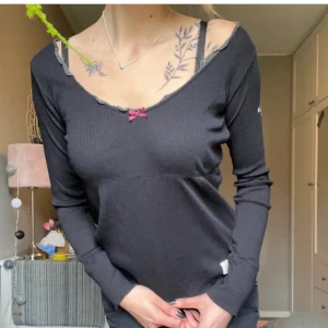 Odd Molly topp  - Säljer en svart långärmad Odd Molly topp med en söt liten rosett framtill. Toppen har en ribbad struktur och en v-ringad design med spetsdetaljer vid halsen. Perfekt för både vardag och festliga tillfällen. Superbekväm och stilren!