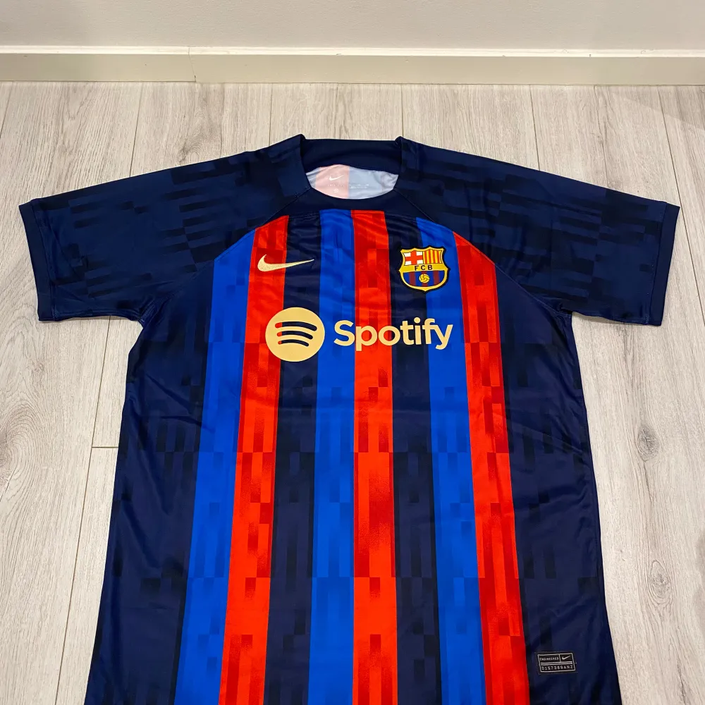 Säljer en FC Barcelona fotbollströja i nyskick. Den är kortärmad och har de klassiska ränderna i rött och blått. Tröjan har sponsorloggor från Spotify och UNHCR. Perfekt för alla Barca-fans! 💙❤️. T-shirts.