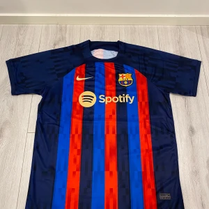 FC Barcelona Fotbollströja - Säljer en FC Barcelona fotbollströja i nyskick. Den är kortärmad och har de klassiska ränderna i rött och blått. Tröjan har sponsorloggor från Spotify och UNHCR. Perfekt för alla Barca-fans! 💙❤️