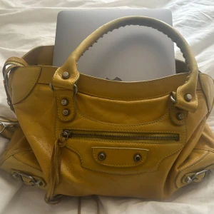 Balenciaga City Bag - Säljer en snygg gul Balenciaga City Bag i skinn. Väskan har en cool design med dragkedja och nitar som . Skinnet har lite rispningar och någon fläck som jag dock inte har testat att tvätta bort,  är i relativt bra skick ändå! Pris kan diskuteras! 