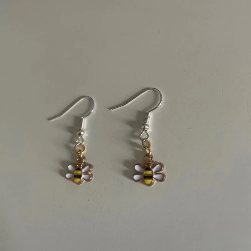 Supergulliga örhängen med små bi-berlocker i gult och svart. De har vita vingar och hänger på en silverfärgad krok. Perfekta för att lägga till en lekfull touch till din outfit. Passar både till vardags och fest!. Asusteet.