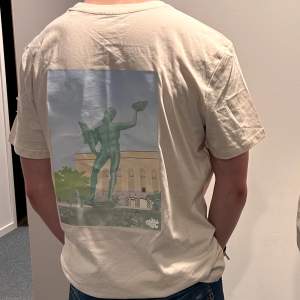 Säljer en vit t-shirt med ett stort tryck på ryggen som är Poseidon framför en byggnad. T-shirten är i bomull och har en normal passform med korta ärmar. Perfekt för sommardagar eller avslappnade tillfällen. Den är 100% miljövänlig och tillverkas av ett UF företag 🌸Mittgöteborg🌸