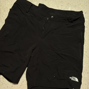 Svarta vandrings shorts från The North Face - Säljer ett par svarta vandrings shorts från The North Face i mycket bra skick. De är perfekta för sommarens äventyr och har en bekväm passform med dragkedja och knapp framtill. Materialet är slitstarkt och passar bra för utomhusaktiviteter. Perfekta för både träning och vardag! Finns små enstaka fläckar från färg men går säkert bort om man andvänder något medel!