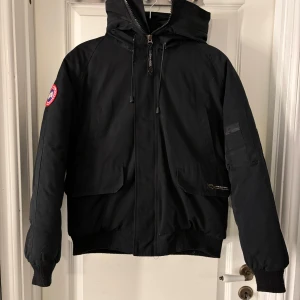Svart jacka från Canada Goose - Säljer en svart jacka från Canada Goose i mycket bra skick. Den har en dragkedja framtill och en huva med snörning. Perfekt för kalla vinterdagar med sin varma och tåliga design. Märkeslogga på ärmen ger en snygg detalj.