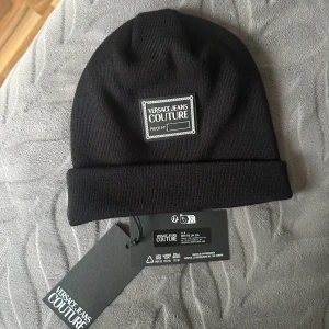 Svart mössa från Versace Jeans Couture - Säljer en snygg svart mössa från Versace Jeans Couture i nytt skick. Den är ribbad och har en stilren design med en broderad logga framtill. Perfekt för att hålla sig varm och trendig under kyliga dagar. Mössan är i one size och passar de flesta.