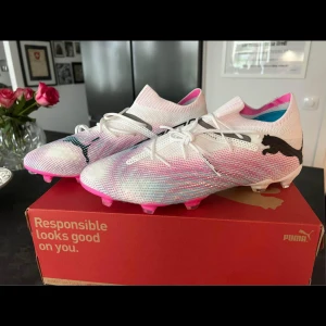 Fotbollsskor Puma Future 7 Ultimate FG/AG - Säljer ett par Puma Future 7 Ultimate FG/AG fotbollsskor i vitt och svart med coola rosa detaljer. Skorna är helt oanvända och perfekta för både gräs och konstgräs. De har en snygg design med snörning och Puma-loggan på sidan. Perfekta för fotbollsspelare som vill sticka ut på planen!
