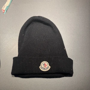 Moncler mössa - Säljer en snygg svart stickad mössa från Moncler. Den är ribbad och har Monclers ikoniska logga framtill. Perfekt för att hålla sig varm under kalla dagar och passar till de flesta outfits. Mössan är i okej skick. Äkta såklart!
