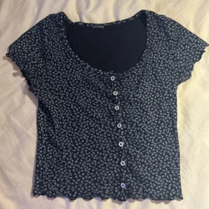 Svart blommig topp med knappar Brandy Melville - Söt svart topp med vita blommor och knappar framtill från Brandy Melville. 