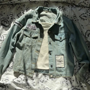 Ljusblå jeansjacka med patchar - Säljer en cool ljusblå jeansjacka med unika patchar och tryck. Jackan har en oversized passform. Perfekt för vår och sommar! Den har knappar framtill och flera detaljer som ger den en unik stil. Passar perfekt för dig som vill sticka ut! Ingår även luva som man knäpper fast 