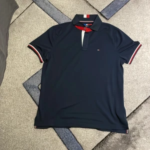 Mörkblå pikétröja från Tommy Hilfiger - Säljer en snygg mörkblå pikétröja från Tommy Hilfiger. Den har en klassisk krage med röd och vit randig detalj och knappar framtill. Ärmarna är korta med matchande ränder i rött och vitt. Perfekt för en stilren och sportig look! Passar även M tajt