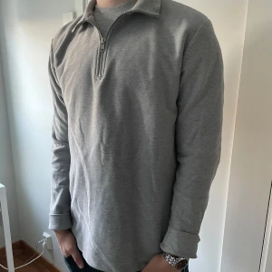 Grå tröja med dragkedja från Topman - Säljer en stilren grå tröja från Topman i storlek M. Den har en praktisk dragkedja vid halsen och är långärmad, perfekt för både vardag och lite kyligare dagar. Materialet känns mjukt och bekvämt. Passar bra till både jeans och chinos!