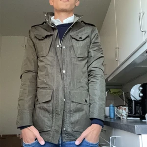 Grön parkajacka med dragkedja - Säljer en snygg grön parkajacka med dragkedja och knappar. Jackan har långa ärmar och flera praktiska fickor framtill. Perfekt för höst och vår!