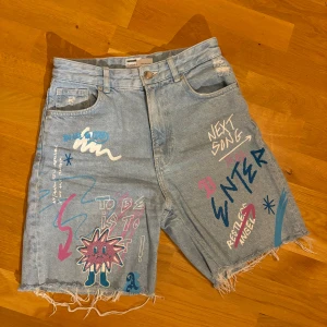 Jeansshorts - Blåa jeansshort med rosa/blå/vita tryck på. Högmidjade och i normal storlek. Storleken 36. Köpta från Bershka.