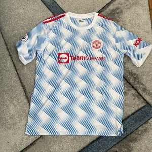 Manchester United fotbollströja - Säljer en snygg Manchester United fotbollströja från Adidas. Den har ett coolt mönster i blått och vitt med röda detaljer. Tröjan är köpt på en streetmarket men är i oanvänt skick då den inte passar min stil