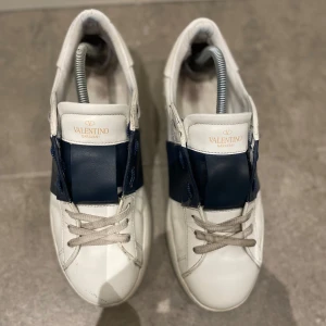 Valentino Garavani sneakers i vitt och blått - Snygga Valentino Garavani sneakers i vitt skinn med en bred blå rem över ovansidan i storlek 41. Skorna har klassisk snörning och en stilren design som passar perfekt för en trendig look. Köparen betalar frakt! 