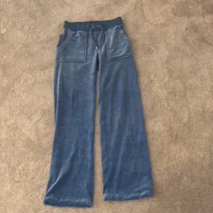 Blå mjukisbyxor från Juicy Couture - Snygga blå mjukisbyxor från Juicy Couture i en supermjuk velour. Byxorna har en bekväm resårmidja med snörning och praktiska fickor både fram och bak. Perfekta för en avslappnad stil med en touch av lyx.