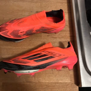 Orange fotbollsskor från Adidas - Säljer ett par skarpa orangea fotbollsskor från Adidas med svarta detaljer och en strömlinjeformad design. Skorna har en lätt och flexibel sula med dobbar för optimalt grepp på planen. Perfekta för den som vill ha både stil och funktion på fotbollsplanen.