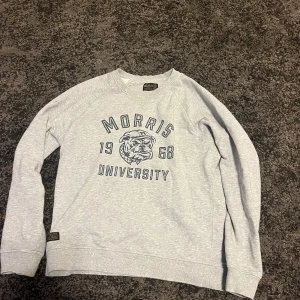 Grå sweatshirt från Morris - Säljer en grå sweatshirt från Morris med tryck av en bulldog och texten 'Morris University 1968'. Tröjan har långa ärmar och en rund halsringning med ribbade muddar vid ärmslut och nederkant. Perfekt för en avslappnad stil.