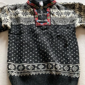Svart och vit stickad tröja med traditionellt mönster från L.M Norway - Säljer en svart och vit stickad tröja med traditionellt norskt mönster. Tröjan har långa ärmar och dekorativa spännen framtill med färgglada detaljer i rött och grönt. Perfekt för kalla dagar!