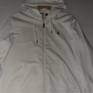 Vit hoodie från Polo Ralph Lauren - Säljer en stilren vit hoodie från Polo Ralph Lauren med dragkedja och justerbar huva. Den har en broderad logotyp på bröstet och praktiska fickor framtill. Perfekt för en avslappnad look.