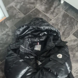 Moncler Maya  - Svart pufferjacka från Moncler. Jackan har en quiltad design med en dragkedja framtill och en huva för extra värme. Den har ett broderat Moncler-märke på ärmen och praktiska fickor med knappar. Har haft den på mig 2-3 gånger. Nypris 7000kr