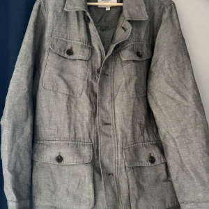 Olivgrön jacka från Dressmann - Snygg grön jacka från Dressmann i linneblandning. Jackan har en klassisk design med knappar och flera praktiska fickor framtill. Perfekt för en stilren look.