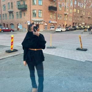 Svart cape  - Snygg svart cape med bälte som ger en elegant look. Perfekt för att hålla sig varm och stilren. Den har en unik design med en asymmetrisk form och är ärmlös, vilket gör den lätt att bära över andra plagg.