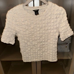 Beige topp från Lindex - Säljer en beige topp från Lindex med korta ärmar och en unik textur. Perfekt för en stilren look. Passar bra till både jeans och kjol.