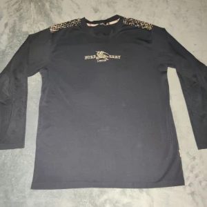 Svart tunn långärmad tröja från Burberry - Säljer en svart och tunn långärmad tröja från Burberry med klassiskt rutmönster på axlarna och Burberry-logga på bröstet. Den är direkt inte helt kol svart, utan en hint av blå. Tröjan är knopprig, använd men väl omhändertagen- små fläckar vid ärmen och på mag delen(se de sista två bilden. Storleken är XL och därför inte passar mig. 100% bomull