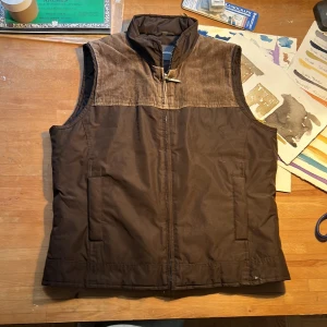 Brun väst från Street One - Säljer en snygg brun väst från Street One. Västen har en quiltad insida och en dragkedja framtill. Den är perfekt för höst och vår med sin stilrena design och bekväma passform. Snygga manchester detaljer. 