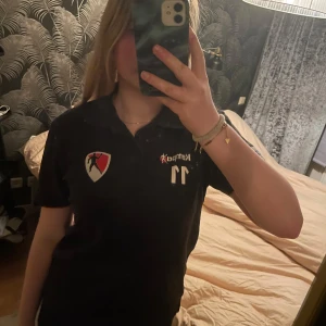 Svart pikétröja från Kempa - Säljer en svart pikétröja från Kempa med nummer 11 tryckt på framsidan. Tröjan har en klassisk krage och korta ärmar. Perfekt för sportiga tillfällen eller som en casual vardagströja. Den har även en logotyp och texten 'Kudos Status' på baksidan.