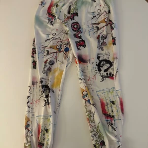 Färgglada byxor med graffiti-mönster - Unika byxor med ett färgglatt graffiti-inspirerat mönster. Basen är vit med inslag av rött, blått, gult och svart. Perfekta för dig som vill sticka ut med en konstnärlig stil.