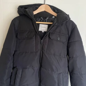 Svart vinterjacka från Jack & Jones - Säljer en svart pufferjacka från Jack & Jones. Jackan har en dragkedja framtill, två bröstfickor med knappar och en huva. Perfekt för kyliga dagar. Storlek xs men passar s.
