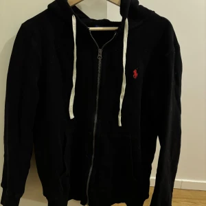 Svart hoodie från Ralph Lauren - Säljer en stilren svart hoodie från Ralph Lauren med dragkedja och vit snörning. Den har en klassisk röd logga på bröstet och är perfekt för en avslappnad look. Passar bra till både jeans och joggers. Använd få gånger.