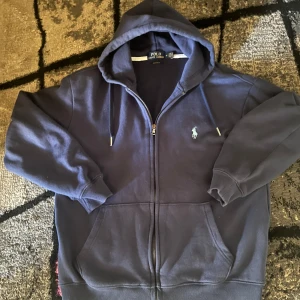 Ralph Lauren zip marinblå - Säljer en snygg blå hoodie från Ralph Lauren med dragkedja och det klassiska logotypbroderiet på bröstet. Perfekt för en avslappnad stil. Hoodien har långa ärmar och en bekväm passform. Denna tröjan är dam storlek så M är egentligen S. Vid några funderingar är det bara att kontakta