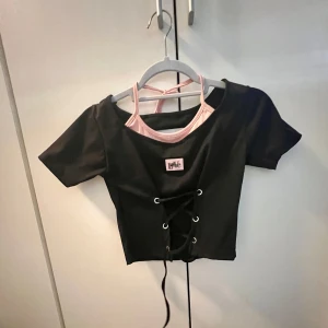 Svart kortärmad topp med snörning - Snygg svart kortärmad topp med rosa detaljer och snörning framtill. Perfekt för en trendig look. Den har en unik design med en rosa insida och ett litet märke på bröstet.
