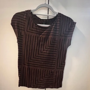 Brun mönstrad topp från Lindex - Säljer en brun topp från Lindex med ett geometriskt mönster i svart. Toppen har korta ärmar och en rund halsringning. Perfekt för en stilren look.