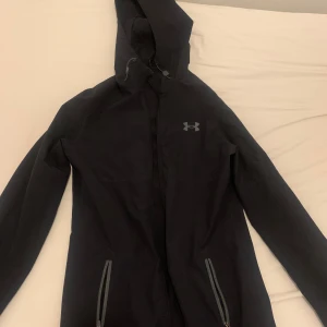 Windbreaker Under Armour dragkedjan är sönder! - Säljer en svart jacka från Under Armour med huva och dragkedja. Perfekt för träning eller vardagsbruk. Jackan har en stilren design med två fickor framtill. 