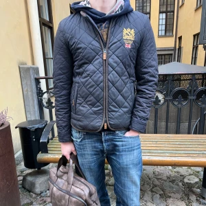 Mörkblå quiltad jacka från Morris - Säljer en snygg mörkblå quiltad jacka från Morris med broderad logga på bröstet. Jackan har dragkedja och långa ärmar, perfekt för höst och vår. Den har en stilren design med ribbade detaljer vid ärmslut och nederkant.