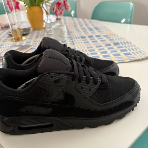 Svarta Nike Air Max 90 - Säljer ett par svarta Nike Air Max sneakers. Skorna har en stilren design med snörning och den klassiska Nike-loggan på hälen. Perfekta för en sportig look.