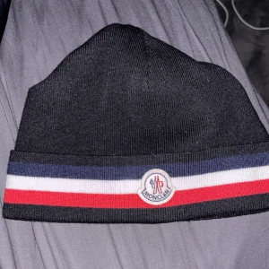 Svart mössa från Moncler - Snygg svart mössa från Moncler med klassisk randig detalj i blått, vitt och rött. Perfekt för kyliga dagar med stil. Märkeslogga framtill ger en exklusiv touch.