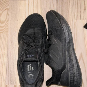 Svarta Adidas löparskor - Säljer ett par svarta Adidas löparskor med snörning och diskret design. Skorna har en bekväm sula och är märkta med 'End Plastic Waste' på insidan. Perfekta för löpning och träning.