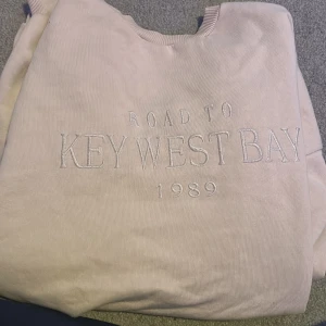 Beige sweatshirt med text - Säljer en beige sweatshirt med texten 'ROAD TO KEY WEST BAY 1989' broderad på framsidan. Tröjan har långa ärmar och ribbade muddar vid ärmslut och nederkant. Perfekt för en avslappnad stil. Stor i storleken 