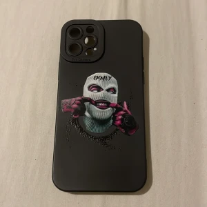 Svart mobilskal med tryck  iPhone 12 Pro  - Snyggt svart mobilskal med ett unikt tryck av en person i rånarluva som gör en grimas. Skalet har en stilren design och skyddar din telefon effektivt. Perfekt för dig som vill ha något annorlunda och coolt.