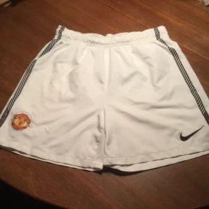 Vita fotbollsshorts från Nike - Säljer ett par vita fotbollsshorts från Nike med Dri-FIT-teknologi. Shortsen har elastisk midja och ett Manchester United-emblem på ena benet. Perfekta för fotbollsträningen eller matchen! ⚽